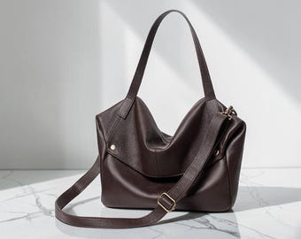 Könnte beinhalten: Eine dunkelbraune Lederschultertasche mit genarbter Textur, die eine lockere Silhouette, einen Tragegriff und einen verstellbaren Umhängeriemen mit goldfarbener Metallschnalle sowie kleinen runden Nieten an den Ecken der Frontklappe aufweist.