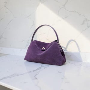 K&ouml;nnte beinhalten: Eine tiefviolette Wildlederhandtasche mit einem einzelnen gebogenen Griff, einem strukturierten Boden und einem silberfarbenen Metalldrehverschluss in der Mitte der Frontklappe. Die Tasche hat eine weiche, matte Textur und eine geschwungene Form.