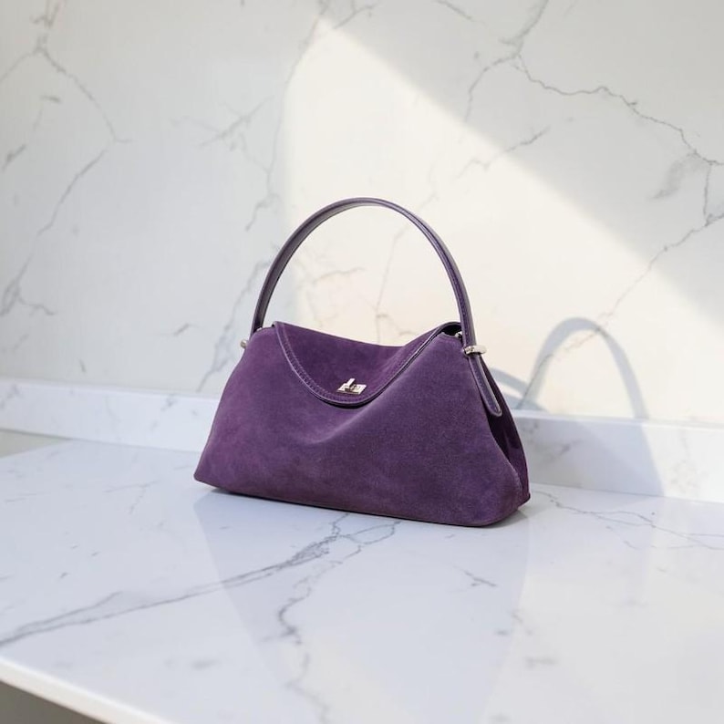 K&ouml;nnte beinhalten: Eine tiefviolette Wildlederhandtasche mit einem einzelnen gebogenen Griff, einem strukturierten Boden und einem silberfarbenen Metalldrehverschluss in der Mitte der Frontklappe. Die Tasche hat eine weiche, matte Textur und eine geschwungene Form.