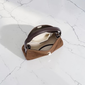 K&ouml;nnte beinhalten: Eine braune Wildlederhandtasche mit einem dunkelbraunen Ledergriff und einem silberfarbenen Metallverschluss. Das Innere ist mit einem hellcremefarbenen Stoff ausgekleidet und verf&uuml;gt &uuml;ber eine offene Tasche, die das offene Design zeigt.