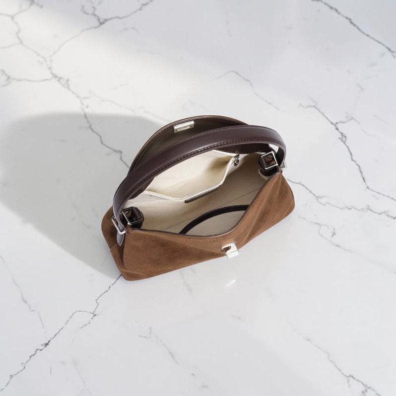 K&ouml;nnte beinhalten: Eine braune Wildlederhandtasche mit einem dunkelbraunen Ledergriff und einem silberfarbenen Metallverschluss. Das Innere ist mit einem hellcremefarbenen Stoff ausgekleidet und verf&uuml;gt &uuml;ber eine offene Tasche, die das offene Design zeigt.