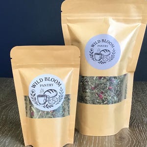 May include: Two brown paper pouches with clear windows, filled with dried herbs and flowers. Each pouch has a circular sticker with the text "Wild Bloom Pantry" and a graphic of a coffee cup, bread, and leaves. The pouches are on a wooden surface.