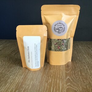 Puede incluir: Dos bolsas de t&eacute; de hierbas de color beige. La bolsa m&aacute;s grande tiene una ventana transparente que muestra la mezcla de t&eacute; y una etiqueta circular que dice "Wild Bloom Pantry". La bolsa m&aacute;s peque&ntilde;a tiene una etiqueta blanca con "Her Radiance Brew".