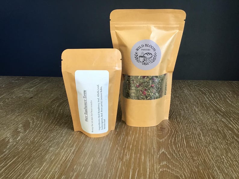Puede incluir: Dos bolsas de t&eacute; de hierbas de color beige. La bolsa m&aacute;s grande tiene una ventana transparente que muestra la mezcla de t&eacute; y una etiqueta circular que dice "Wild Bloom Pantry". La bolsa m&aacute;s peque&ntilde;a tiene una etiqueta blanca con "Her Radiance Brew".