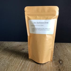 Puede incluir: Una bolsa de papel marr&oacute;n de t&eacute; "Her Radiance Brew". La etiqueta incluye instrucciones de preparaci&oacute;n y enumera los ingredientes: hoja de frambuesa roja, hoja de alfalfa, hierba de lim&oacute;n, p&eacute;talo de rosa roja, melisa y hoja de ortiga.