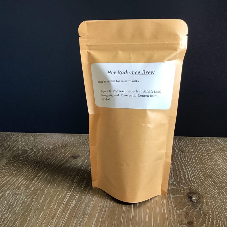 Puede incluir: Una bolsa de papel marr&oacute;n de t&eacute; "Her Radiance Brew". La etiqueta incluye instrucciones de preparaci&oacute;n y enumera los ingredientes: hoja de frambuesa roja, hoja de alfalfa, hierba de lim&oacute;n, p&eacute;talo de rosa roja, melisa y hoja de ortiga.