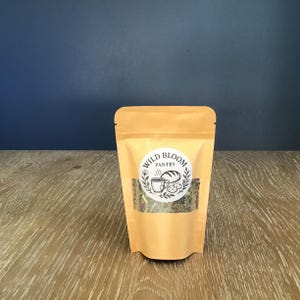 Puede incluir: Una bolsa de color beige con una ventana transparente, llena de hierbas y especias secas. La bolsa tiene una etiqueta circular con el texto "Wild Bloom Pantry" y una ilustraci&oacute;n de una taza, pan y hojas. El fondo es una superficie de madera y una pared azul oscuro.