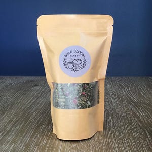 Puede incluir: Una bolsa de papel kraft con una ventana transparente que muestra una mezcla de hierbas y flores secas. La bolsa tiene una etiqueta circular con el texto "Wild Bloom Pantry" e ilustraciones.