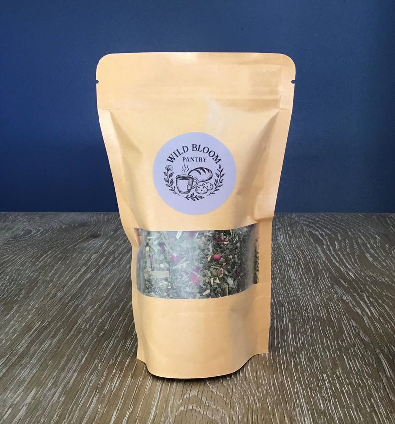 Puede incluir: Una bolsa de papel kraft con una ventana transparente que muestra una mezcla de hierbas y flores secas. La bolsa tiene una etiqueta circular con el texto "Wild Bloom Pantry" e ilustraciones.