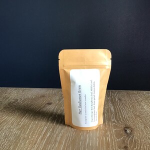 Puede incluir: Una bolsa de papel kraft con una etiqueta blanca que dice "Her Radiance Brew". La etiqueta enumera los ingredientes y las instrucciones de infusi&oacute;n. La bolsa est&aacute; sobre una superficie de madera con un fondo azul oscuro.