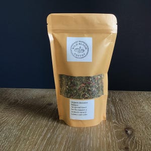 Puede incluir: Una bolsa de papel marr&oacute;n con una ventana transparente que muestra una mezcla de hierbas y flores. La bolsa tiene una etiqueta blanca con el texto "Wild Bloom Pantry" y una etiqueta m&aacute;s peque&ntilde;a que dice "Organic Hormone Balance". Sobre una superficie de madera.