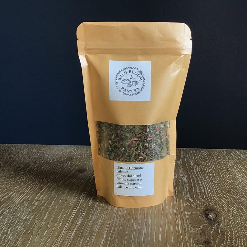Puede incluir: Una bolsa de papel marr&oacute;n con una ventana transparente que muestra una mezcla de hierbas y flores. La bolsa tiene una etiqueta blanca con el texto "Wild Bloom Pantry" y una etiqueta m&aacute;s peque&ntilde;a que dice "Organic Hormone Balance". Sobre una superficie de madera.