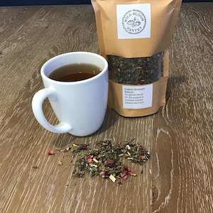 Puede incluir: Una taza de cer&aacute;mica blanca llena de t&eacute; est&aacute; junto a una bolsa de papel marr&oacute;n de t&eacute; de hojas sueltas. La bolsa tiene una etiqueta que dice "Wild Bloom Pantry" y "Organic Hormone Balance". Un peque&ntilde;o mont&oacute;n de t&eacute; suelto y p&eacute;talos de rosa est&aacute; esparcido sobre la superficie de madera.