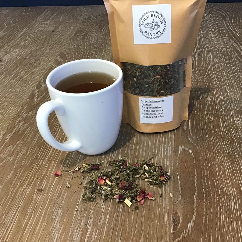 Puede incluir: Una taza de cer&aacute;mica blanca llena de t&eacute; est&aacute; junto a una bolsa de papel marr&oacute;n de t&eacute; de hojas sueltas. La bolsa tiene una etiqueta que dice "Wild Bloom Pantry" y "Organic Hormone Balance". Un peque&ntilde;o mont&oacute;n de t&eacute; suelto y p&eacute;talos de rosa est&aacute; esparcido sobre la superficie de madera.