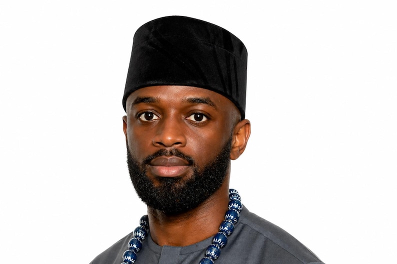 Puede incluir: Retrato de un hombre con una gorra de terciopelo negro y una camisa gris. Tiene barba completa y un collar de cuentas con cuentas azules y blancas. El fondo es blanco liso.