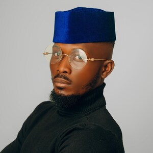 Puede incluir: Un hombre con una gorra de terciopelo azul y gafas con montura dorada. Tambi&eacute;n lleva un cuello alto negro. La imagen est&aacute; tomada sobre un fondo gris. El logotipo "CARL-WINSTON" est&aacute; en la esquina inferior derecha.