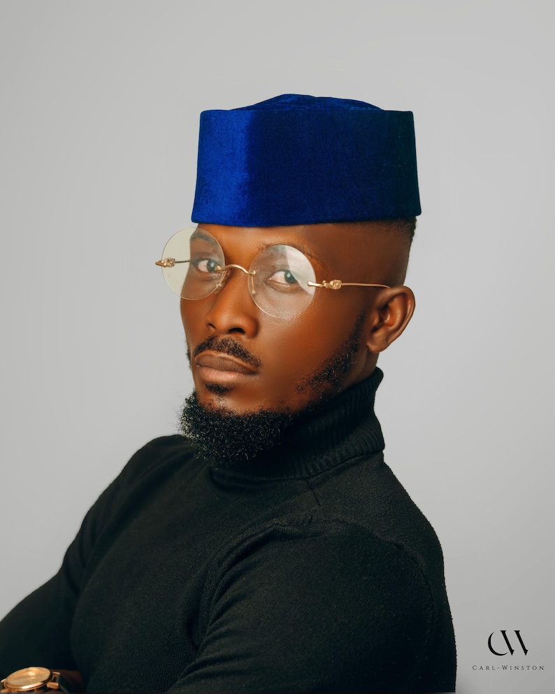 Puede incluir: Un hombre con una gorra de terciopelo azul y gafas con montura dorada. Tambi&eacute;n lleva un cuello alto negro. La imagen est&aacute; tomada sobre un fondo gris. El logotipo "CARL-WINSTON" est&aacute; en la esquina inferior derecha.