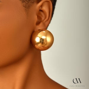 May include: A large, spherical gold earring with a smooth, polished, and reflective surface. The earring is circular in shape and features a high-shine metallic finish. The text 'CW CARL-WINSTON' is visible in the bottom right corner of the image.