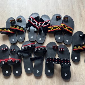 May include: A collection of black sandals with various strap designs. Some straps feature red, white, and yellow embroidered patterns. The sandals are arranged on a light-colored wooden surface, showcasing a variety of sizes.