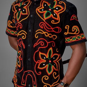Può includere: Camicia nera a maniche corte con ricami floreali colorati. La camicia ha una chiusura con bottoni e un colletto. Il ricamo include dettagli rossi, gialli e verdi. La camicia è realizzata in un materiale morbido, simile al velluto.