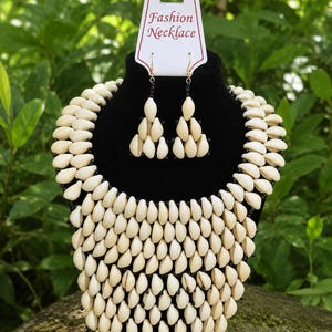 May include: A cream-colored shell necklace and matching earrings set displayed on a black mannequin. The necklace and earrings are made of small, teardrop-shaped shells. The earrings have black beads and gold-colored hooks. The tag reads "Fashion Necklace."