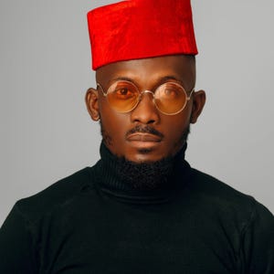 Puede incluir: Retrato de una persona con sombrero rojo, gafas con montura dorada y lentes naranjas, y un jersey de cuello alto negro. La persona tiene barba y bigote bien recortados. El fondo es gris liso.