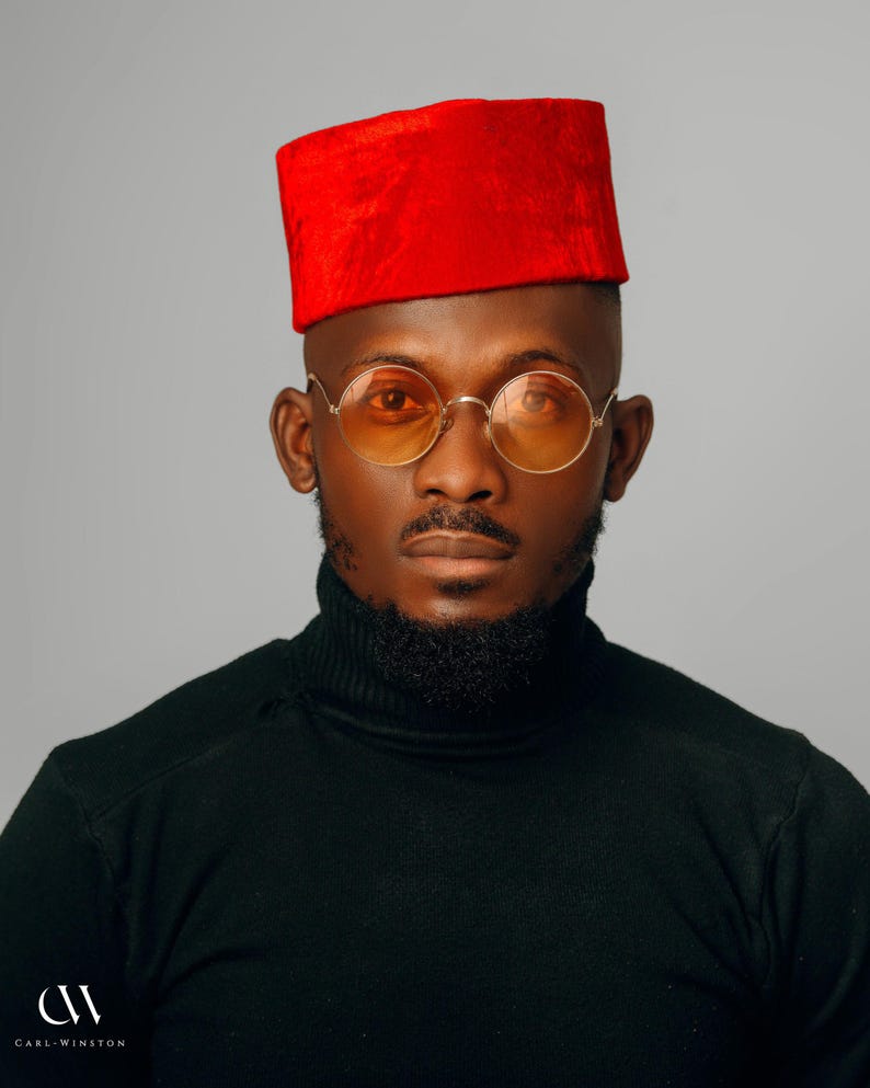 Puede incluir: Retrato de una persona con sombrero rojo, gafas con montura dorada y lentes naranjas, y un jersey de cuello alto negro. La persona tiene barba y bigote bien recortados. El fondo es gris liso.