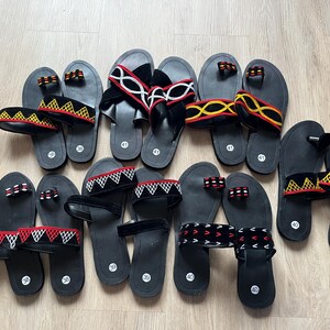 K&ouml;nnte beinhalten: Eine Auswahl an schwarzen Sandalen mit verschiedenen Riemen. Einige Riemen weisen geometrische Muster in Rot, Wei&szlig; und Gelb auf, andere sind einfarbig schwarz oder wei&szlig;. Die Sandalen liegen auf einer hellen Holzoberfl&auml;che.