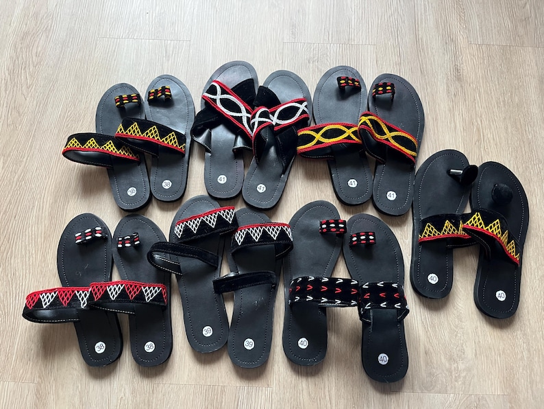 K&ouml;nnte beinhalten: Eine Auswahl an schwarzen Sandalen mit verschiedenen Riemen. Einige Riemen weisen geometrische Muster in Rot, Wei&szlig; und Gelb auf, andere sind einfarbig schwarz oder wei&szlig;. Die Sandalen liegen auf einer hellen Holzoberfl&auml;che.