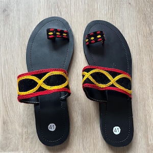 K&ouml;nnte beinhalten: Ein Paar schwarze Sandalen mit dekorativen Riemen. Die Sandalen haben eine Zehenschlaufe und einen Riemen &uuml;ber dem Fu&szlig;, beide mit rot, gelb und schwarz gestickten Mustern. Die Sohlen sind mit der Nummer 41 markiert.