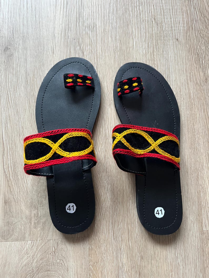 K&ouml;nnte beinhalten: Ein Paar schwarze Sandalen mit dekorativen Riemen. Die Sandalen haben eine Zehenschlaufe und einen Riemen &uuml;ber dem Fu&szlig;, beide mit rot, gelb und schwarz gestickten Mustern. Die Sohlen sind mit der Nummer 41 markiert.