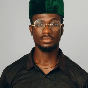 Puede incluir: Un hombre con un gorro kufi de terciopelo verde oscuro, gafas redondas con montura dorada y una camisa negra estampada. Se ve un collar de cadena dorada. La camisa tiene el logo "CW CARL-WINSTON" en la parte inferior izquierda.
