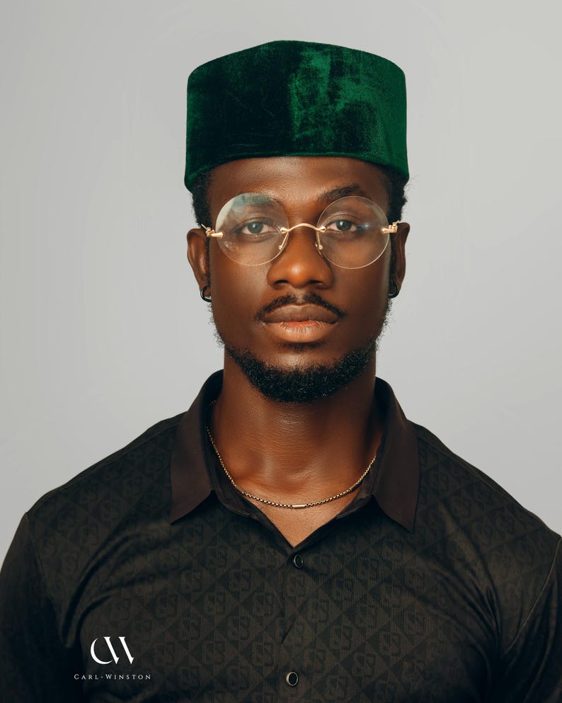 Puede incluir: Un hombre con un gorro kufi de terciopelo verde oscuro, gafas redondas con montura dorada y una camisa negra estampada. Se ve un collar de cadena dorada. La camisa tiene el logo "CW CARL-WINSTON" en la parte inferior izquierda.