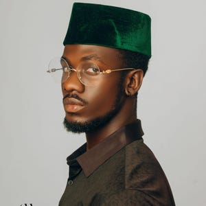 Puede incluir: Una persona con un fez de terciopelo verde oscuro y gafas redondas con montura dorada. La persona lleva una camisa marr&oacute;n oscuro estampada con cuello marr&oacute;n. La imagen incluye el texto "Carl Winston".