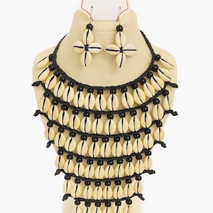 May include: A cream-colored shell necklace and matching earrings set with black accents. The necklace features multiple strands of shells and black beads. The earrings are four-petal flower shaped. The tag reads "Fashion Necklace".