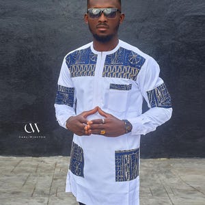 May include: A white long-sleeved shirt featuring blue and gold patterned detailing on the shoulders, chest, and pockets. The shirt has a mandarin collar and a front zip. The brand name "Carl-Winston" is visible.