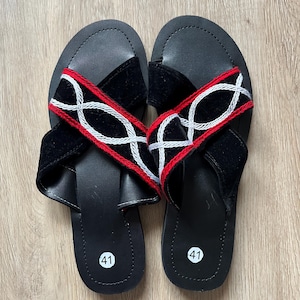 K&ouml;nnte beinhalten: Ein Paar schwarze Ledersandalen mit Kreuzriemen-Design. Die Riemen sind aus schwarzem Samt mit roten und wei&szlig;en Stickereien. Die Sandalen haben eine Gr&ouml;&szlig;enangabe von 41.