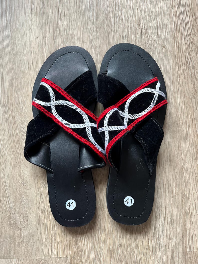K&ouml;nnte beinhalten: Ein Paar schwarze Ledersandalen mit Kreuzriemen-Design. Die Riemen sind aus schwarzem Samt mit roten und wei&szlig;en Stickereien. Die Sandalen haben eine Gr&ouml;&szlig;enangabe von 41.