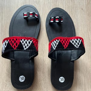 May include: Pair of black leather sandals with red, white, and black embroidered straps. The sandals have a toe loop and a strap across the top of the foot. The soles are marked with the number 38.