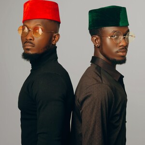Puede incluir: Dos hombres con gafas redondas con montura dorada y sombreros a juego. Un sombrero es rojo, el otro verde. Uno lleva un cuello alto negro, el otro una camisa marr&oacute;n estampada. El fondo es gris liso.