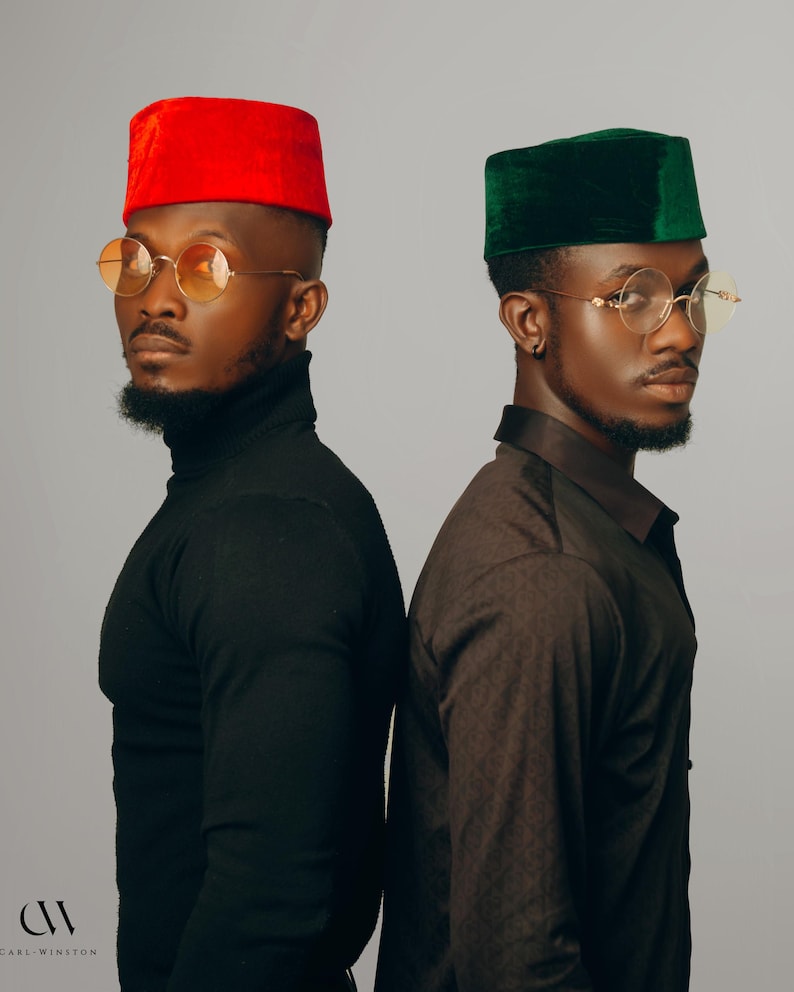 Puede incluir: Dos hombres con gafas redondas con montura dorada y sombreros a juego. Un sombrero es rojo, el otro verde. Uno lleva un cuello alto negro, el otro una camisa marr&oacute;n estampada. El fondo es gris liso.
