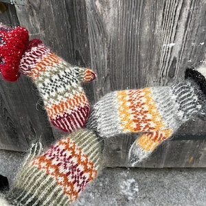 May include: A group of knitted mittens in various colours. The mittens display detailed patterns in red, orange, green, grey, and white. They are made from a soft, textured yarn, ideal for cold weather.