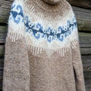 Op de afbeelding: Een warme, handgebreide trui met een coltrui. De trui heeft een colorblock-ontwerp met tinten bruin, crème en blauw. De blauwe accenten zijn in een decoratief patroon over de borst en op de manchetten.