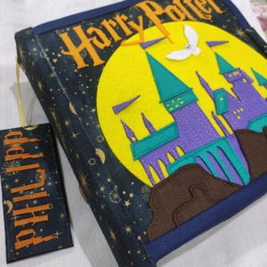 Op de afbeelding: Een handgemaakte Harry Potter boekomslag met bijpassende bladwijzer. De omslag is marineblauw met gouden sterren en de woorden "Harry Potter" in goud. Een vilten kasteelontwerp in paars, teal en geel staat op de voorkant. De bladwijzer heeft de naam "Philipp" in oranje.