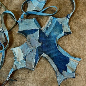 May include: A patchwork denim accessory with a unique, asymmetrical shape. The item is constructed from various shades of blue denim, stitched together with visible white thread. The design includes long straps, suggesting it may be a bag or garment.