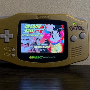 Puede incluir: Consola Game Boy Advance dorada que muestra un juego de Dragon Ball GT. La pantalla muestra un personaje con un atuendo rojo y rosa. La consola tiene una cruceta, botones de inicio, selección, A y B. La palabra "Pokemon" también es visible.