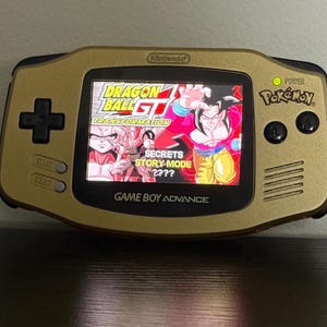 Puede incluir: Consola portátil Game Boy Advance dorada con pantalla mostrando un juego de Dragon Ball GT. La pantalla muestra personajes en acción. La consola tiene una cruceta, botones de inicio y selección, y botones de encendido y Pokemon.