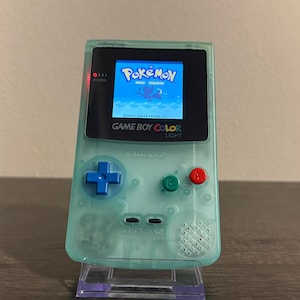 以下が含まれることがあります： 半透明のミントグリーンの携帯ゲーム機。バックライト付きの画面には「Pokémon Gold Version」のタイトル画面が表示されています。青い十字キー、緑のBボタン、赤いAボタン、黒いセレクトボタンとスタートボタンが配置されています。