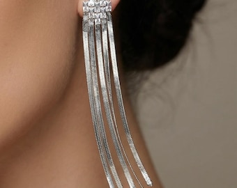 May include: Silver-coloured dangle earrings. The earrings have a geometric top with multiple dangling strands. The earrings are worn on a person's ear, with the face and shoulder visible.