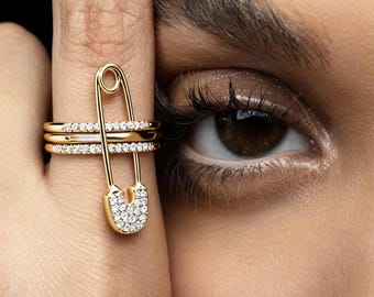Könnte beinhalten: Ein goldfarbener Sicherheitsnadelring mit einer Reihe kleiner, klarer Steine am unteren Ende. Der Ring befindet sich an einem Finger, mit zwei zusätzlichen goldenen Ringen mit klaren Steinen. Der Hintergrund ist schwarz.